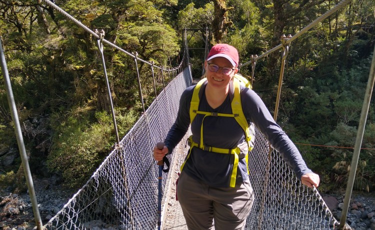 A person wearing a red cap, dark top, and yellow backpack stands on a swing bridge.