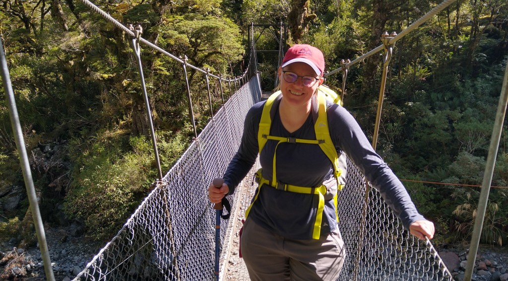 Deborah on a bridge on the Milford Track. She sears a red cap, a dark top, and a yellow back pack.