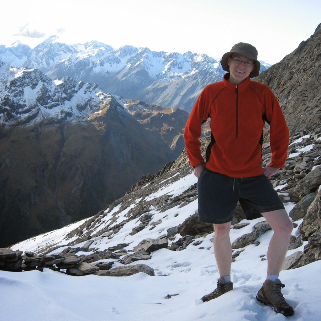 Deborah wears an orange jumper and blue shorts. She is standing in snow with mountains in the background.