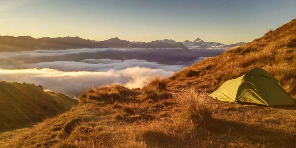 Golden sunlight streams over the scene. A light green tent is pitched on golden tussock. The background is a layer of cloud and mountains.