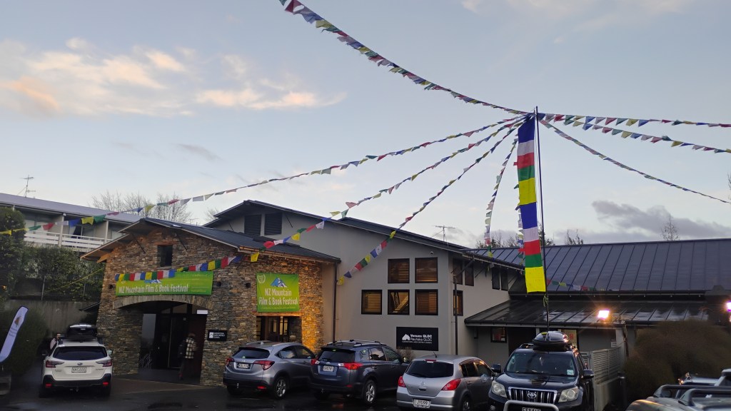 The Wanaka Center building. It has a stone entrance facade and a grey main building. From a tent pole hang Tibetan prayer flags.