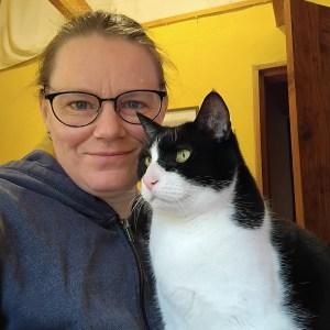 Deborah is in her office with her cat. The office walls are yellow. She smiles at the camera. The cat is a black and white tuxedo, with a pink nose and olive green eyes. She's looking out the window.