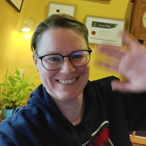 Deborah smiles and waves at the camera. She has dark, roundish glasses on, and a dark blue hoodie.