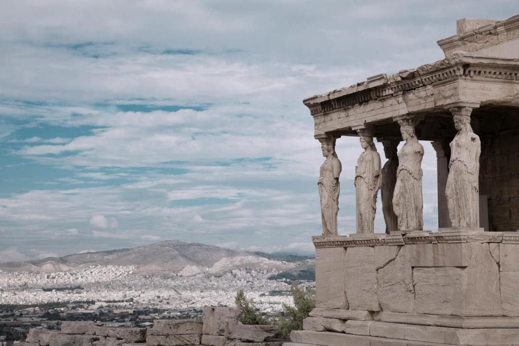 In the foreground, caryatids holding up the Erechtheion in Athens, Greece. In the background, a view of Athens.