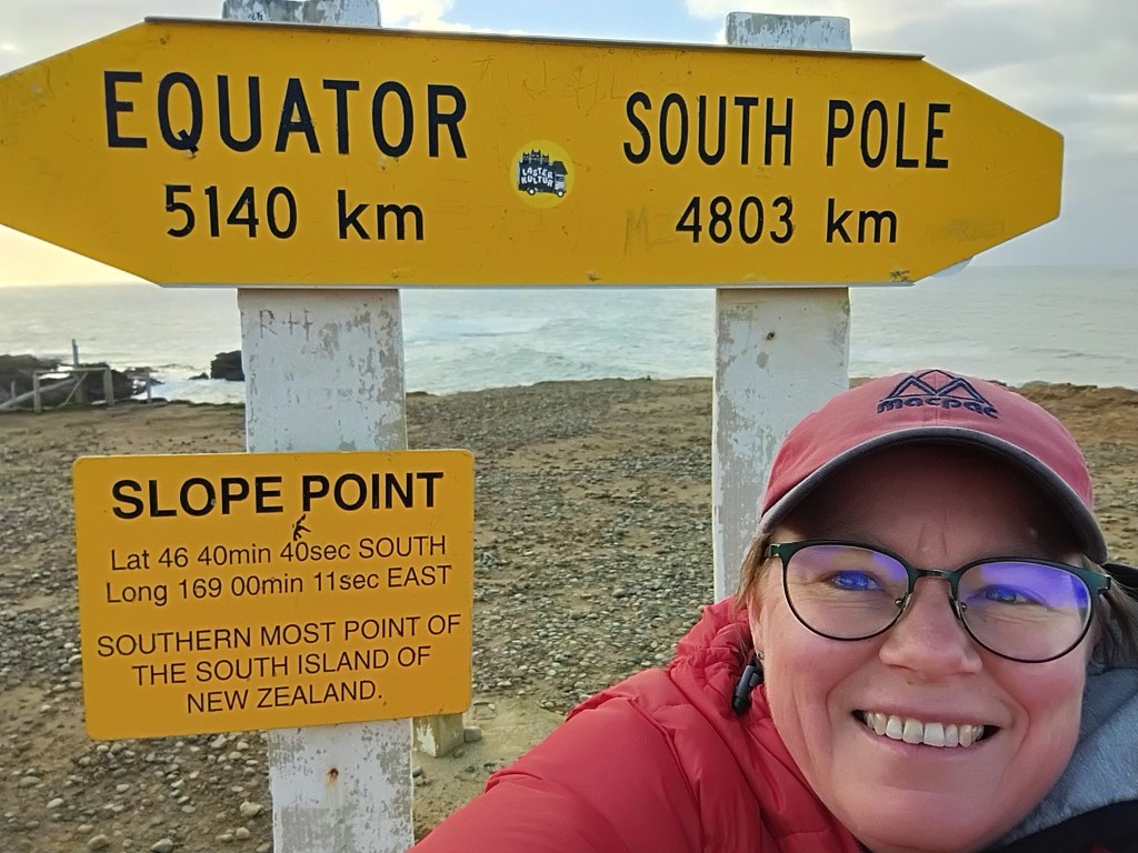 A selfie of Deborah with the Slope Point sign. The left edge points to the equator and gives a distance of 5140 kilometres. The right edge points to the South Pole and says 4803 kilometres.