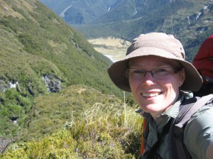Deborah smiles at the camera. She is hearing a tan bucket hat and light green shirt. The valley falls away in the background.