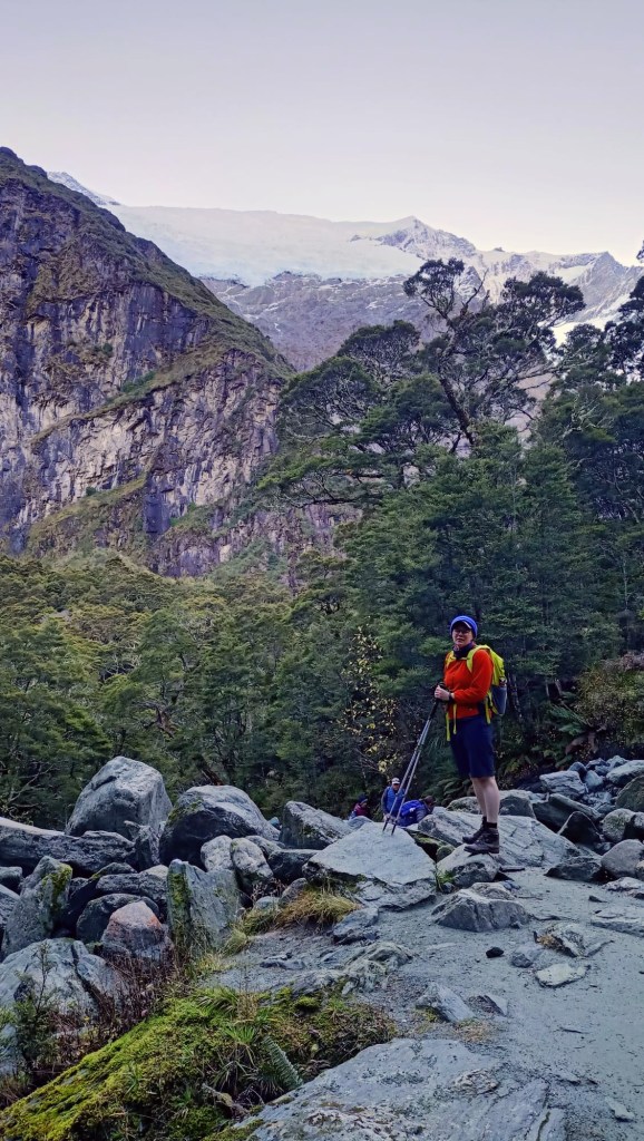 Deborah stands in the foreground wearing an orange jumper and blue shorts. In the mid-ground is rocks and beech trees, In the background is the Rob Roy Glacier clinging to the side of the mountain.